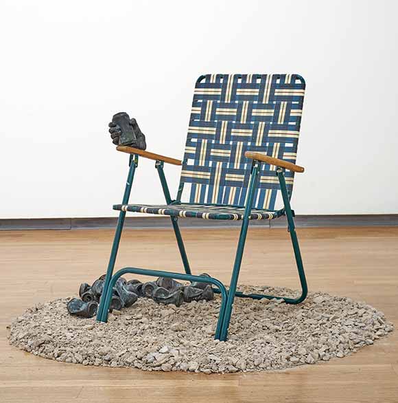 A sculpture of a lawn chair with a disembodied hand clutching a can of beer, with many others crumbled below on the floor.