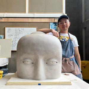 An artist in a blue cap and blue apron leaning on a ceramic sculpture that is half a giant head