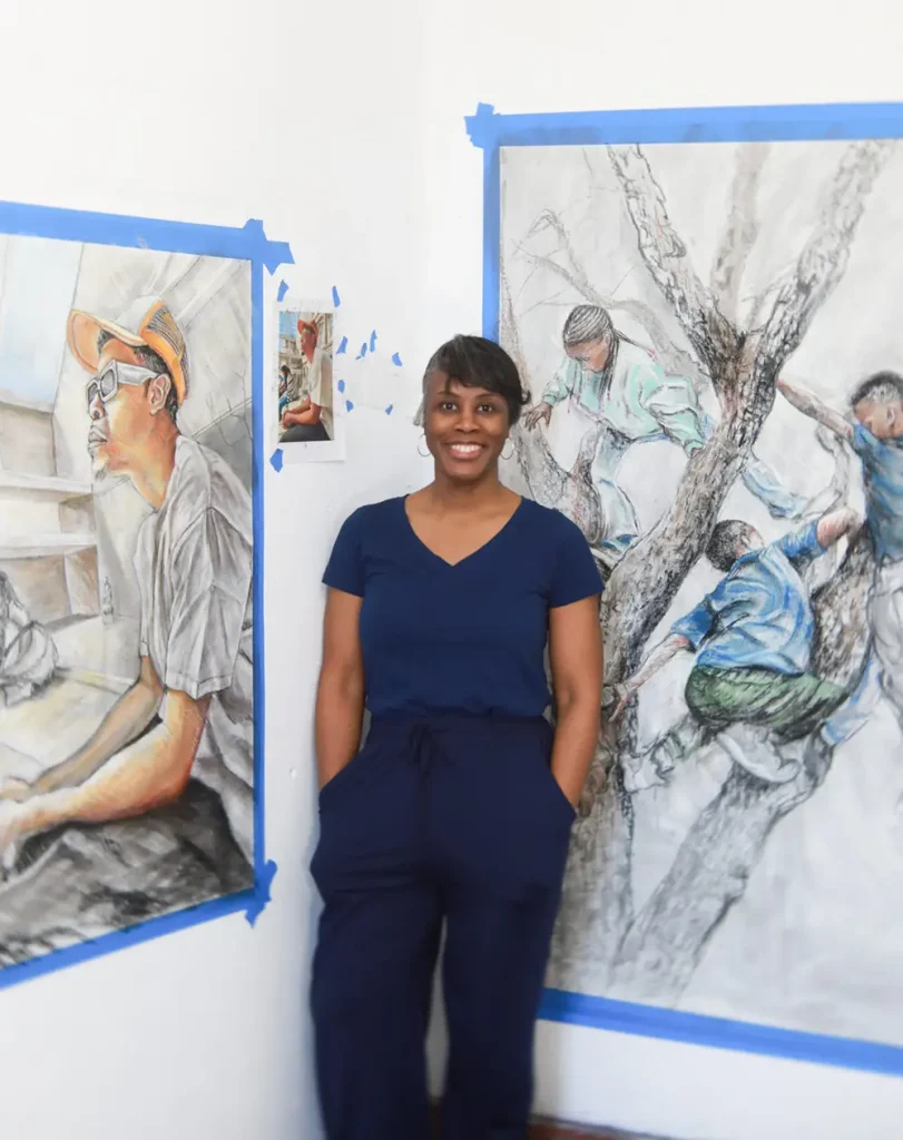 Sukenya Best stands in front of two of her artwork drawings, one of which depicts children climbing a tree. 