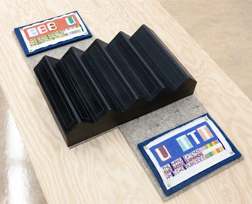 A piece of artwork is displaye dagainst a wooden table. The artwork appears to be a carved piece of steel, wool, and felt. 