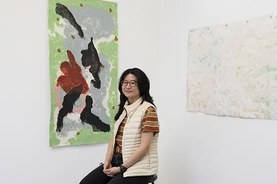 Danqi Cai sits in front of two of her artwork pieces that hang on the wall behind her. 