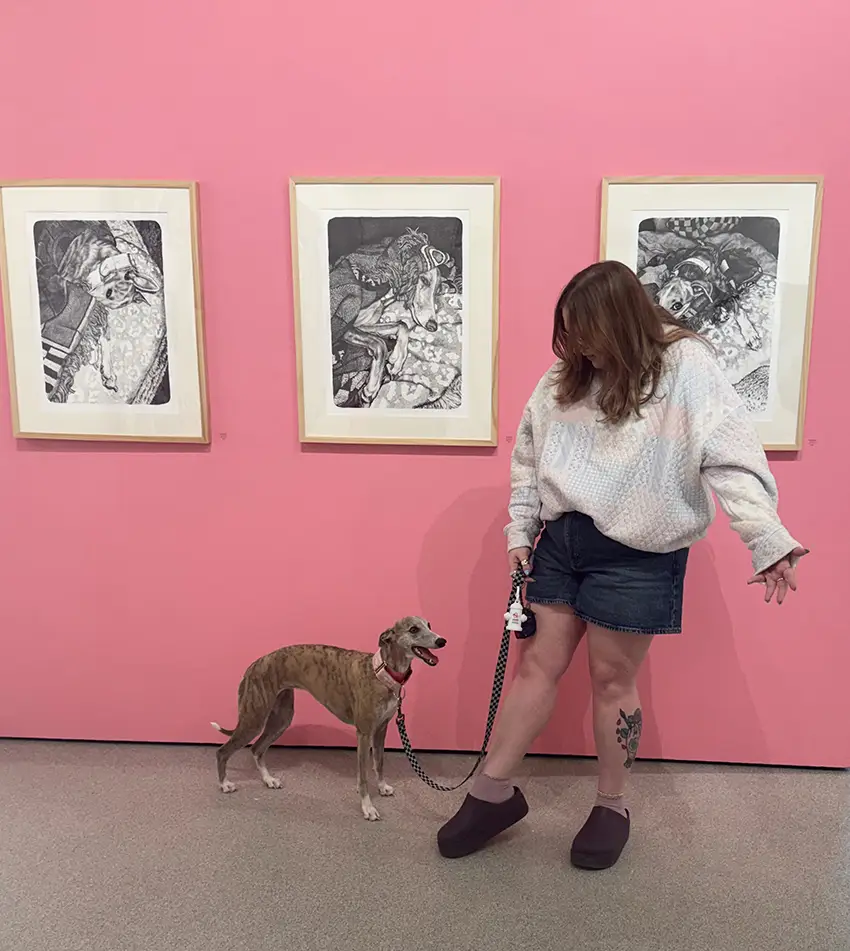 Shannon Ferguson stands in front of three wall art pieces hanging against a pink painted wall. Her dog stands next to her. 