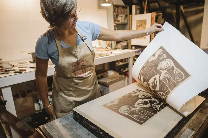 Lee Marchalonis holds a large printed book open while wearing an apron and working at her letterpress. 