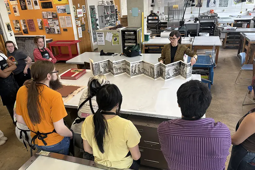 A professor shows an accordion book to a group of students while working in the Art Printshop.